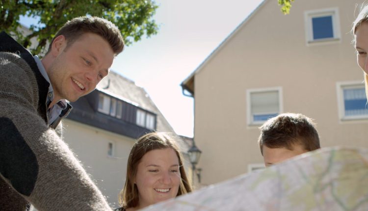Vier Personen werden aus der Froschperspektive gezeigt, wie sie auf eine Landkarte schauen. Im Hintergrund ist eine Hausfassade zu erkennen.