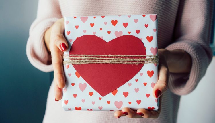 Weibliche Hände halten ein Geschenk, eingewickelt in Herzpapier.